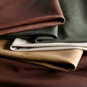 Cortina Leathers Linden full grain collection hero photo shows naked aniline leather hides in rich browns flanking a moss green, a pearl white and a buttery yellow, showcased with deep shadows to highlight the pure aniline tonal variation and natural markings.